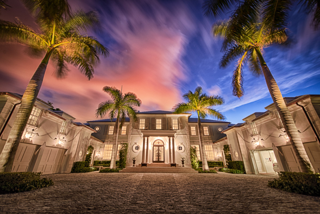 Grand palm-lined estate glows at dusk, its arched entry and windows lit beneath a pink and blue streaked sky.