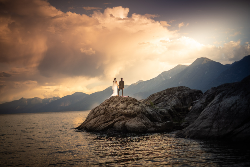 A wedding couple stands hand in hand on a rocky point above Kootenay Lake, BC, beneath a glowing sunset sky.