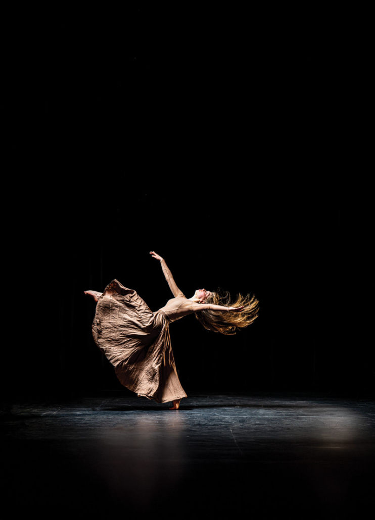A dancer arches backward mid-spin, flowing dress and hair caught in a single spotlight on a dark stage.