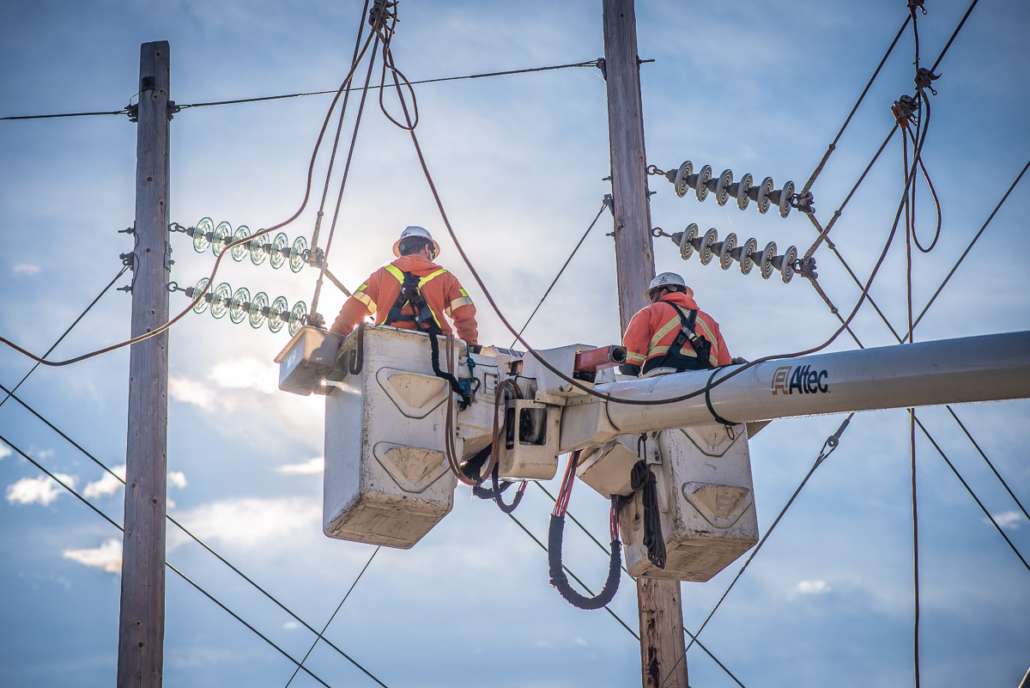 Two linemen in bucket trucks work on high-voltage power lines against a bright sky.