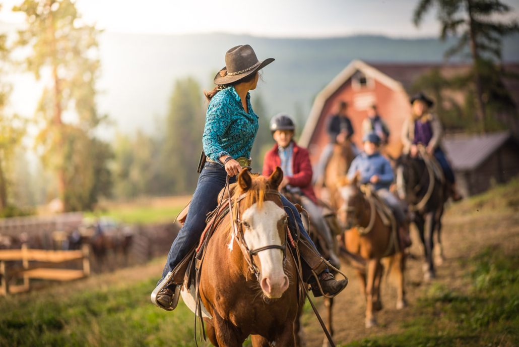 A guide leads riders on horseback past a red barn at a Cranbrook, BC ranch in golden morning light.