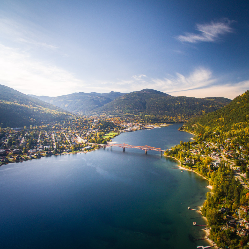 Aerial view of an orange bridge spanning a lake between a forested mountain town and tree-lined shoreline.