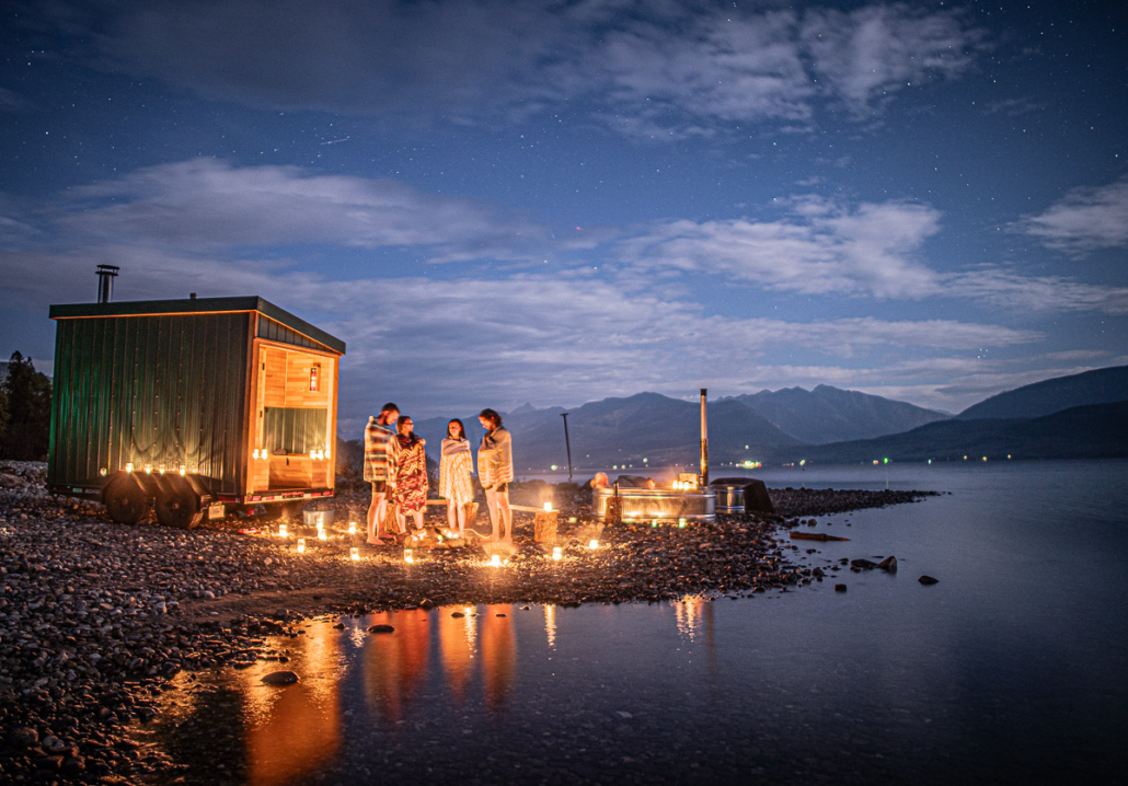 Wrapped in towels, four people gather by a lakeside sauna and candlelit hot tub under a starry dusk sky.