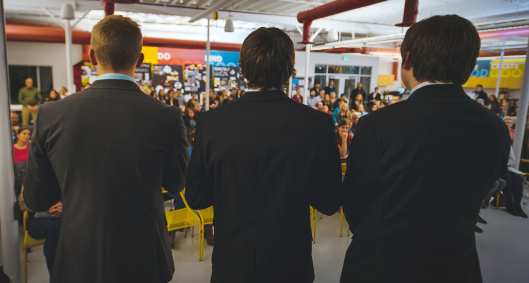 Three men in dark suits face a seated crowd in a bright industrial-style venue with painted slogans on the walls.