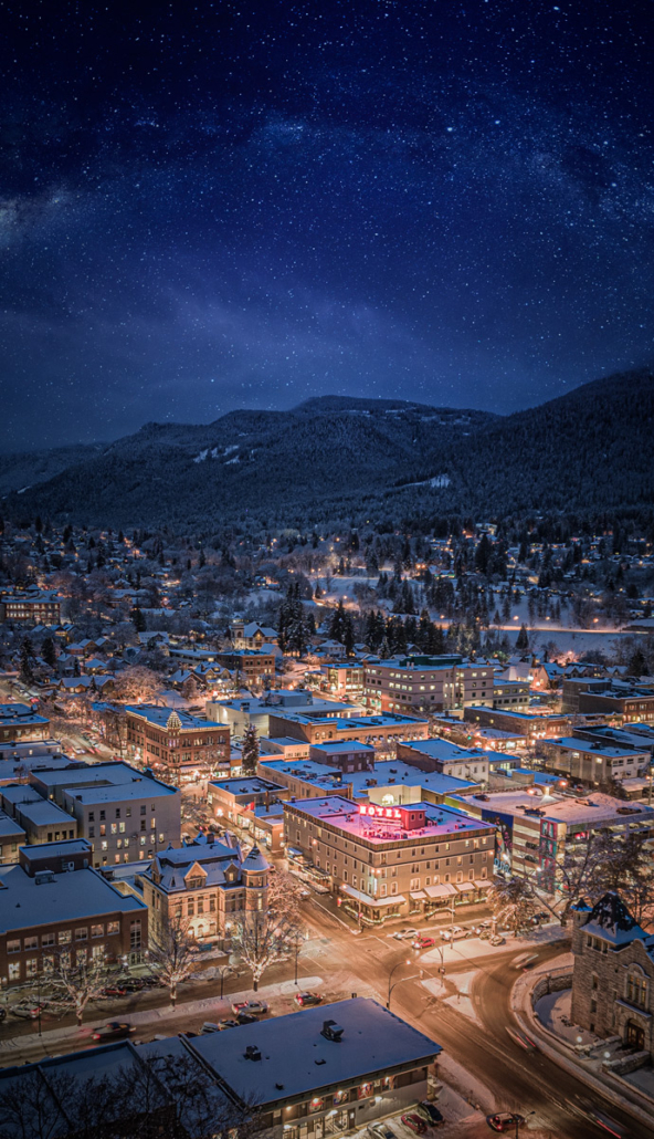Aerial night view of snowy downtown Nelson, BC, with the hotel's neon sign glowing beneath a starry sky.