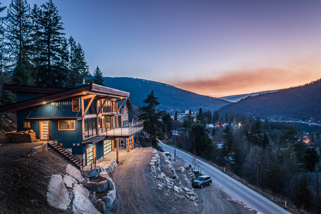 A modern timber-frame home glows at dusk on a hillside above Nelson, BC, with mountains beyond.