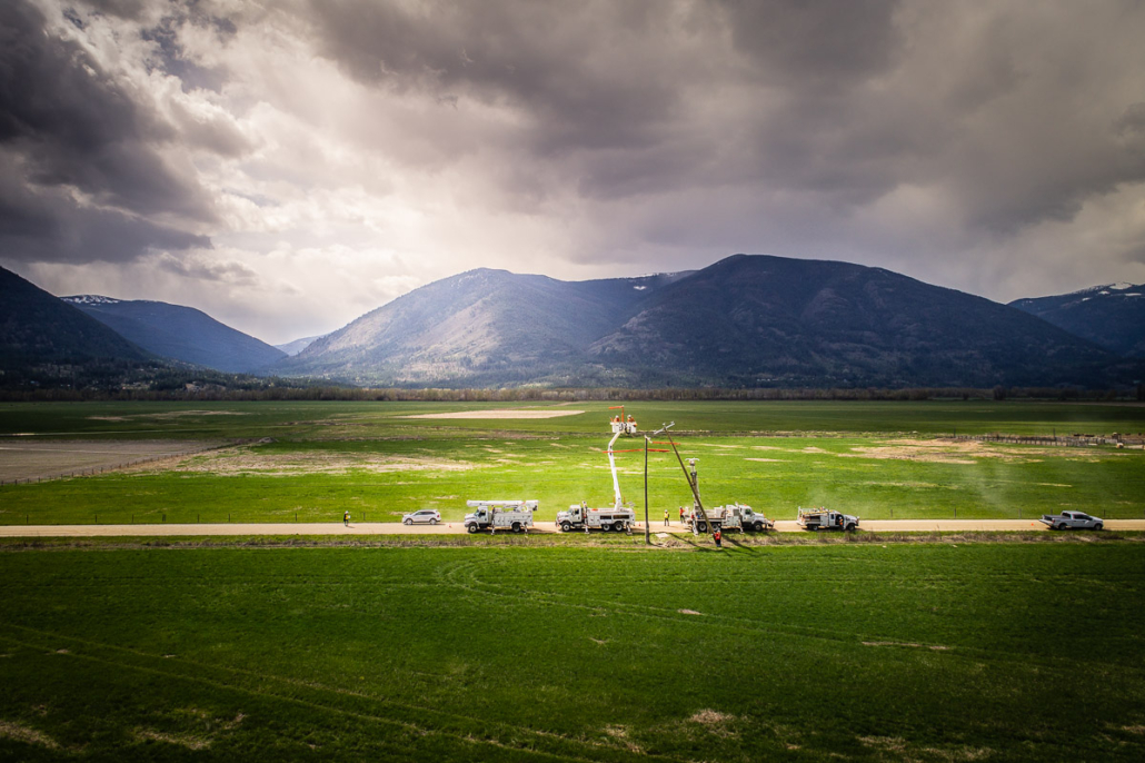 Bucket trucks work on power lines beside a green Kootenay valley field under dramatic storm clouds and mountains.
