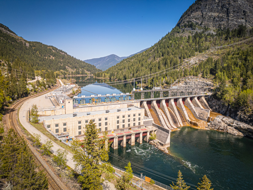 Aerial view of a concrete hydroelectric dam and powerhouse spanning a forested valley river.