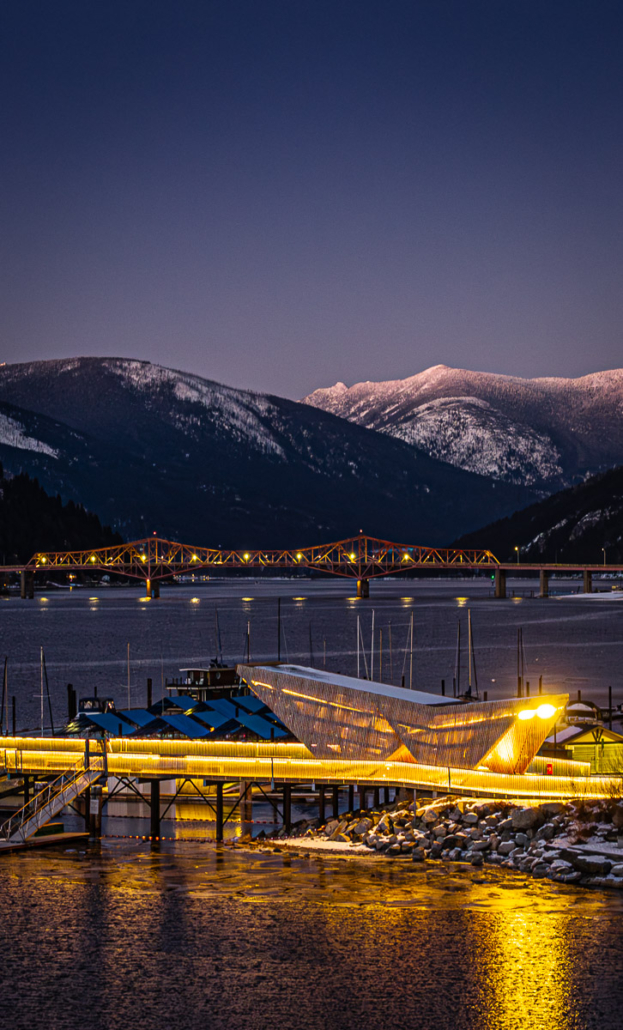 Golden lights outline a lakeside marina and bridge in Nelson, BC, with snowy Kootenay peaks at dusk.