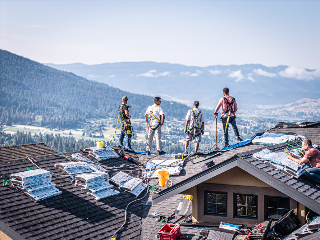 Roofers in safety harnesses install shingles atop a home overlooking a valley near Vernon, BC.