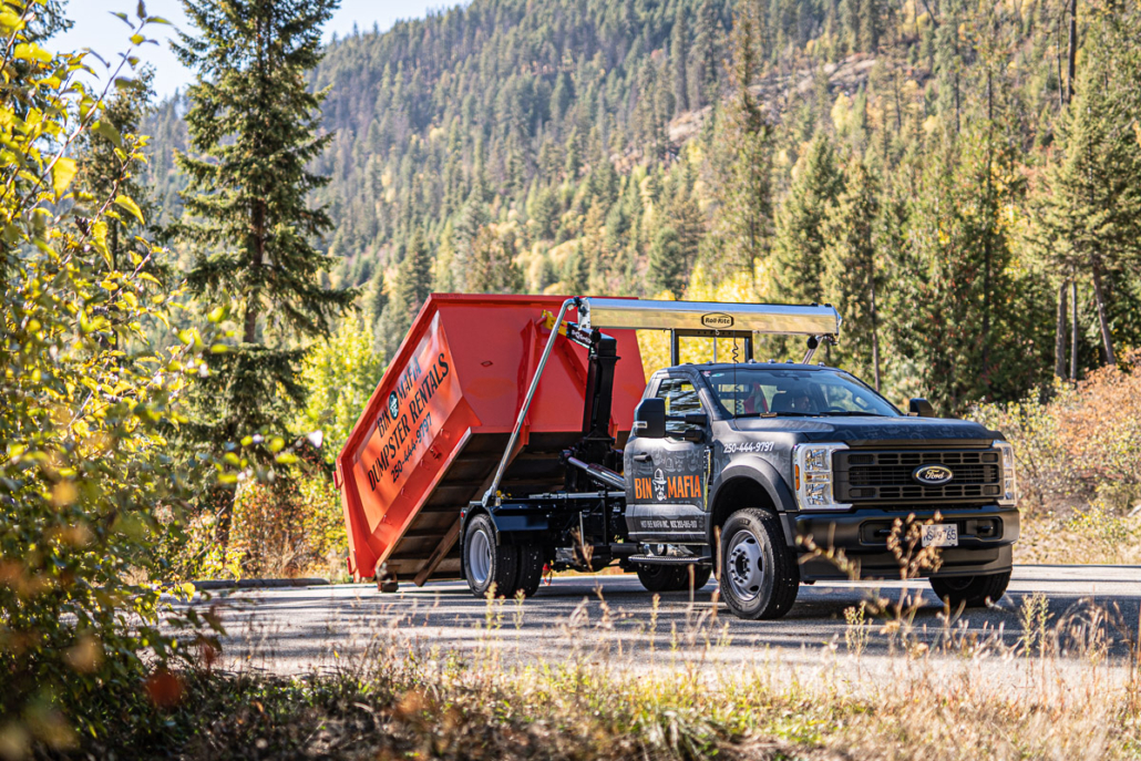 A dumpster rental truck with a tilted orange bin parked on a forested Kootenay roadside near Christina Lake, BC.