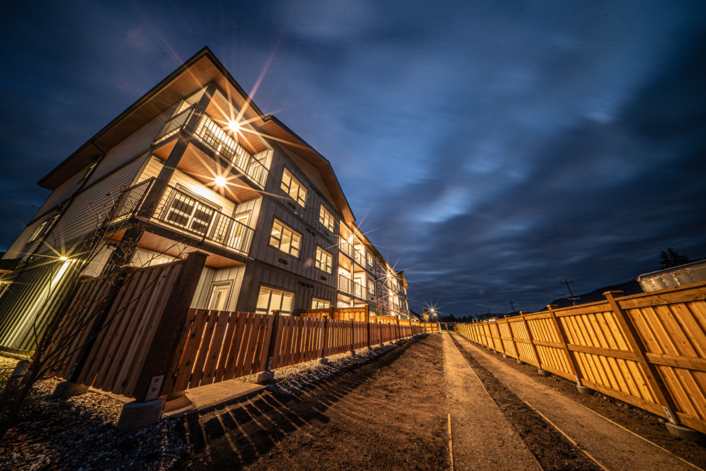Twilight view of a Nelson, BC multi-unit building with glowing balcony lights beside a new wood fence.