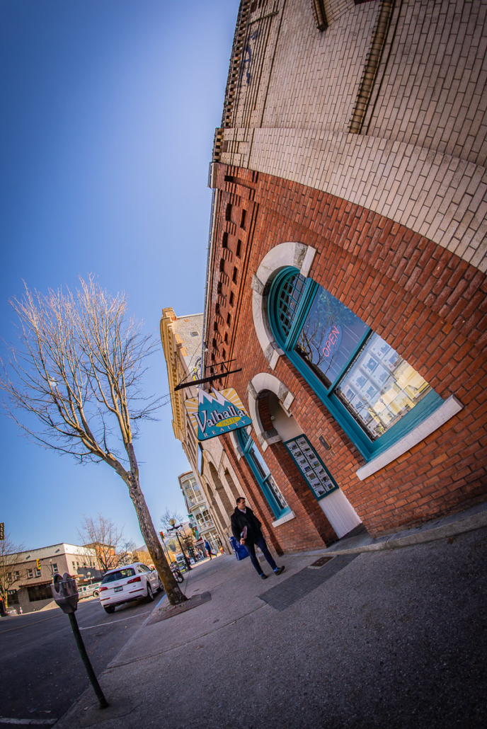 Wide-angle view of a brick heritage building with teal-trimmed windows and a hanging Valhalla Path Realty sign in Nelson, BC.