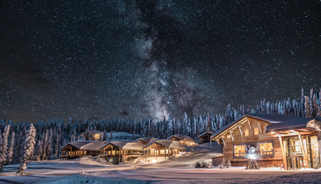 Snow-covered lodge buildings glow with warm light under a starry sky and visible Milky Way.
