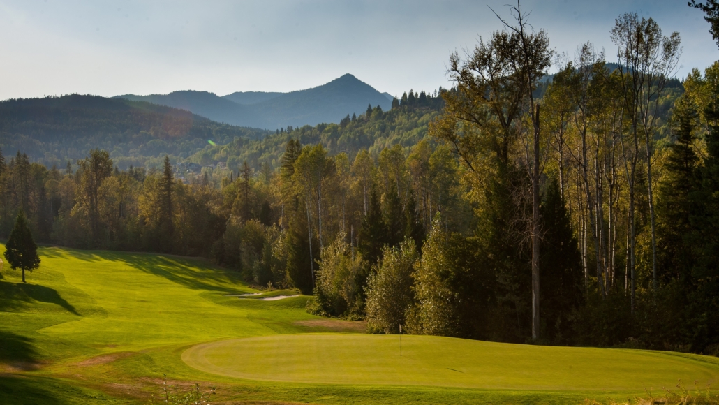 A golf green bordered by tall forest, with Kootenay mountain peaks rising in the hazy distance near Rossland, BC.