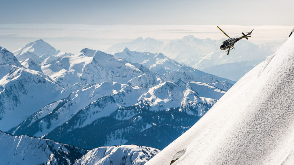 A helicopter flies over snow-capped Kootenay peaks near Revelstoke, ready for a backcountry ski landing.