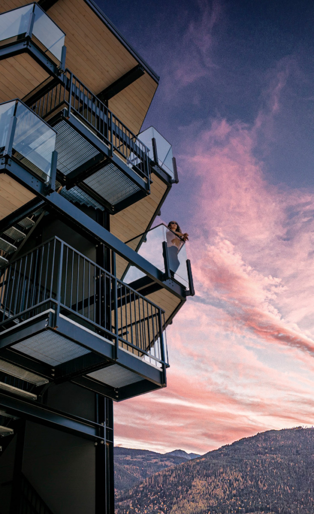 A woman leans over a glass balcony railing at the Stirling Hotel, pink sunset clouds over Kootenay mountains.