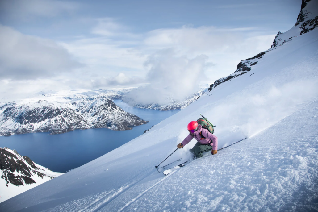 Skier in a pink jacket carves down a steep snowy slope above a deep blue fjord and mountains.