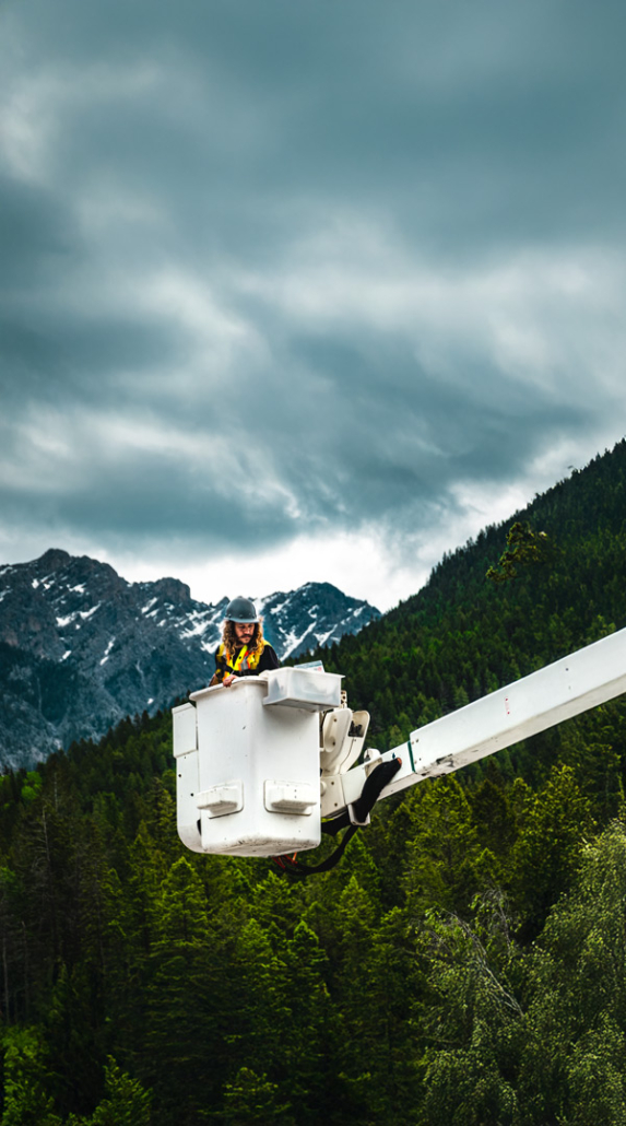 A worker in a hard hat rides a bucket truck lift high above forested Kootenay mountains under stormy clouds.
