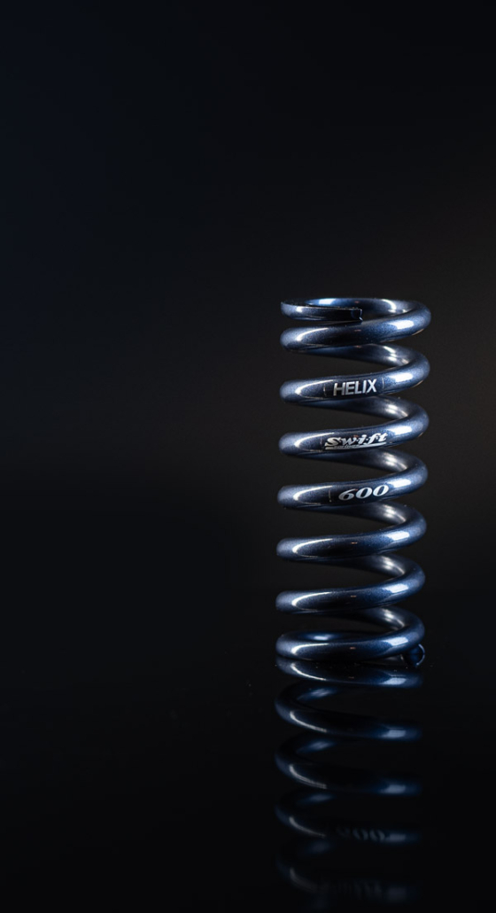 A blue Helix suspension coil spring labelled Swift 600, lit against a black background in Nelson, BC.